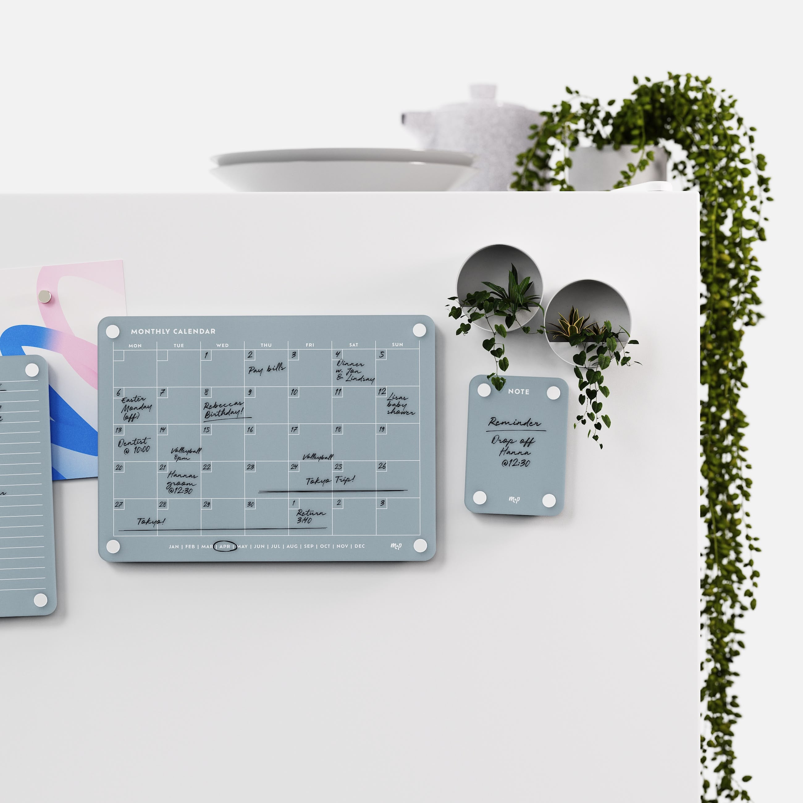 Slate grey monthly calendar and note board on a white kitchen wall with magnetic planters and trailing ivy
